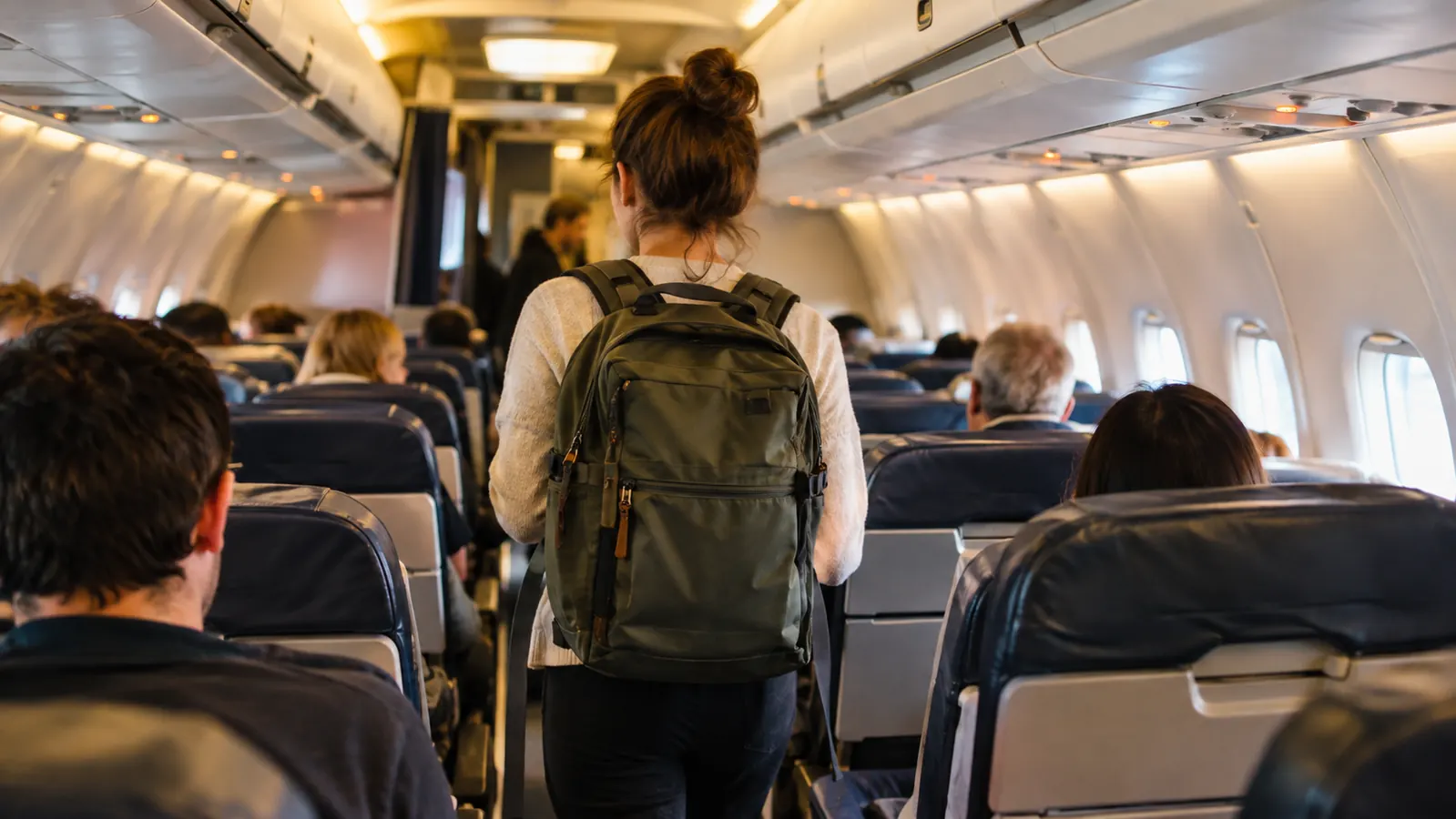 A traveler wearing a travel backpack walks down a narrow airplane aisle toward a window seat