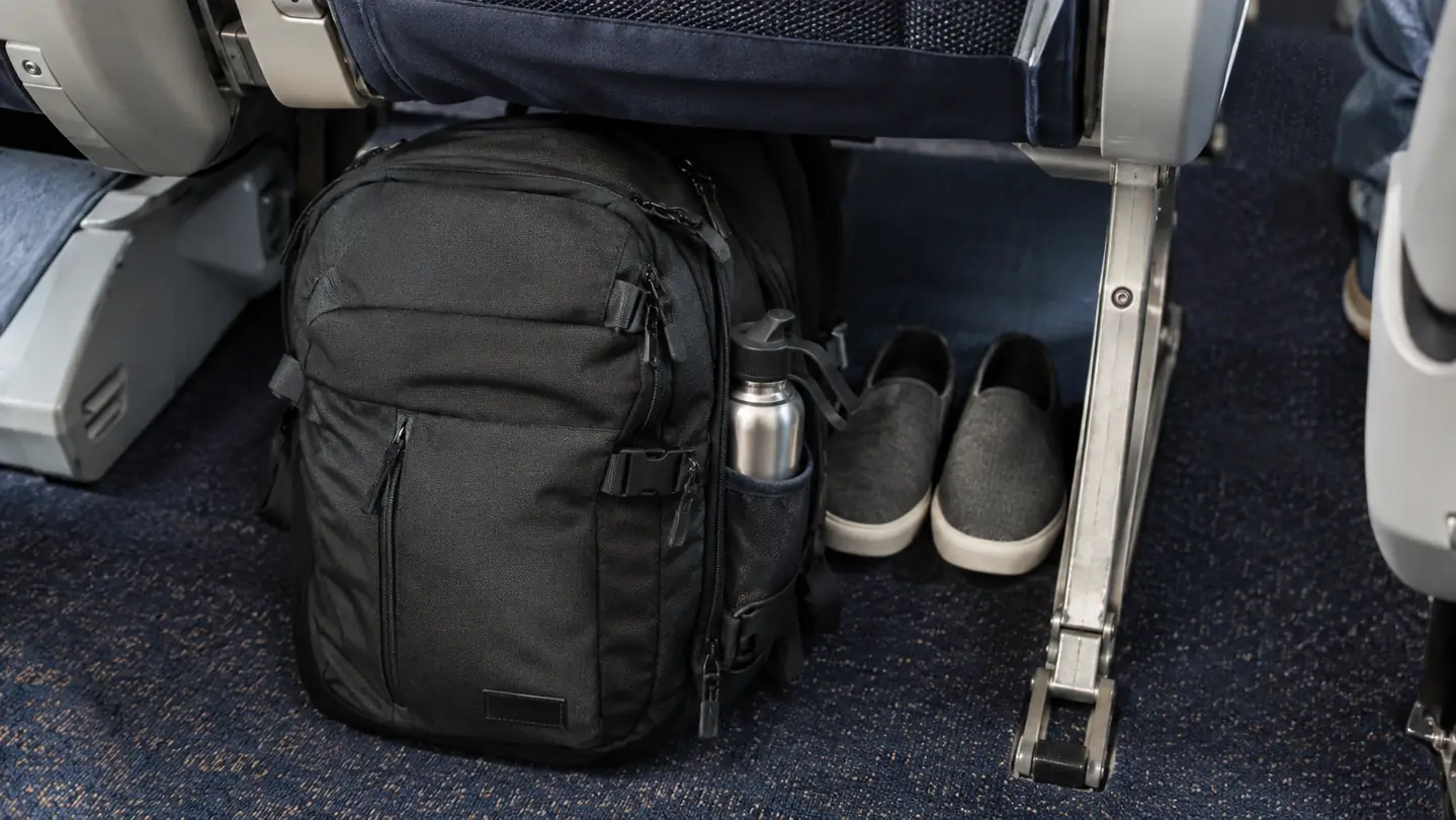 A travel backpack standing upright under an airplane seat with a water bottle in the side pocket