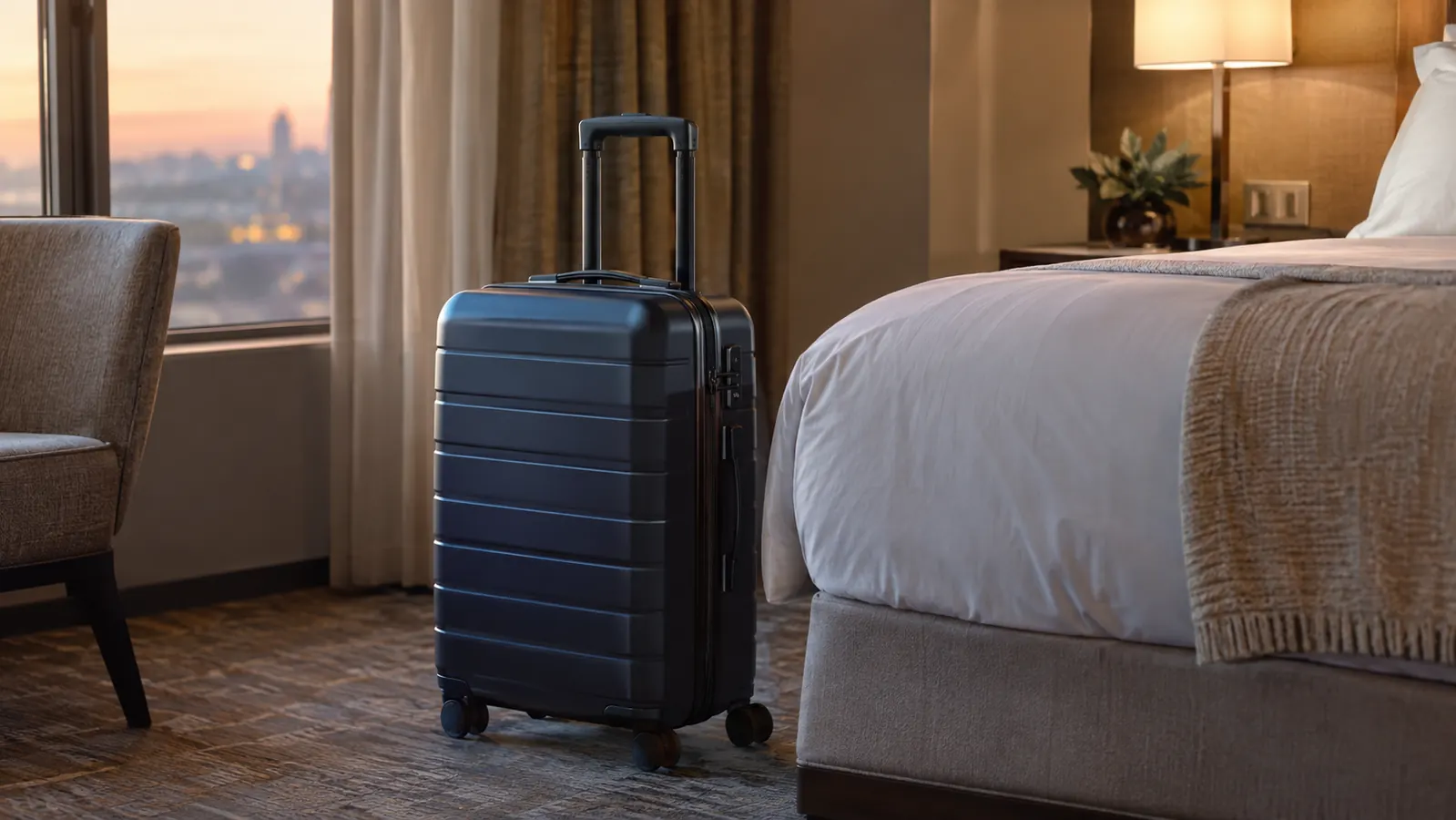A hardside carry-on suitcase standing upright next to a hotel bed with a window behind it